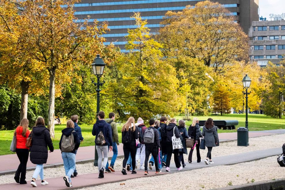 Een groep scholieren die een educatief programma van ProDemos volgen lopen buiten. Op de achtergrond zie je bomen in herfstkleuren en het station Den Haag CS.