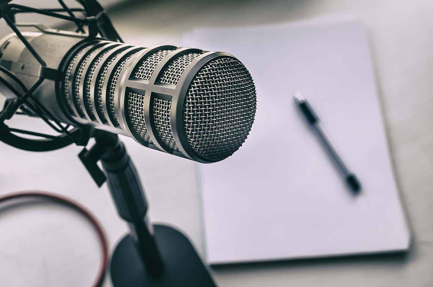 Een professionele microfoon in zilvergrijs. Op de achtergrond ligt een zwarte pen op een wit vel papier op een grijze tafel.