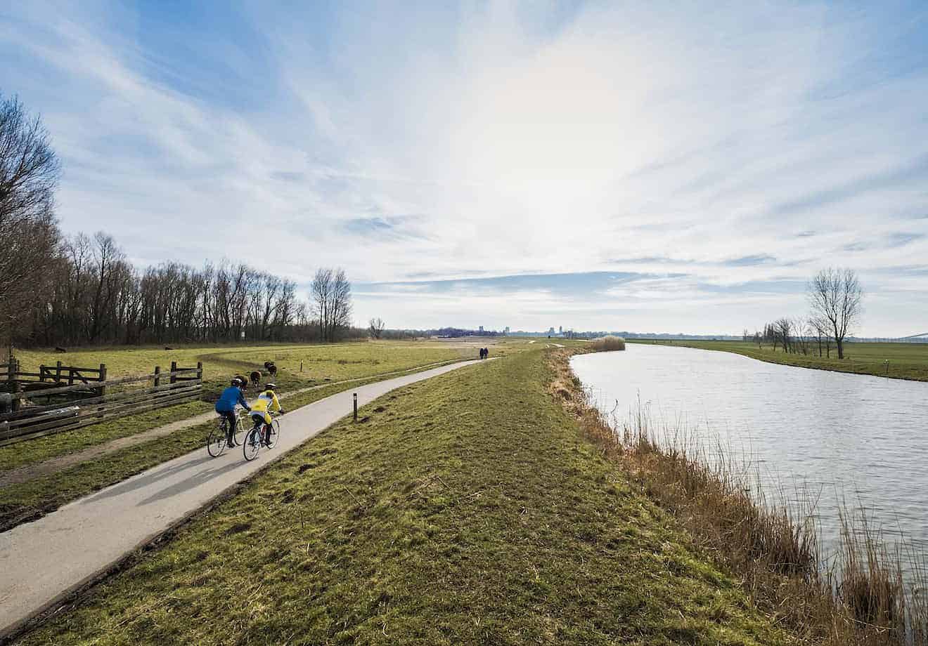 Twee wielrenners fietsen op een weg langs een groene strook gras en een kanaal richting de horizon. De lucht is blauwe met witte wolken.