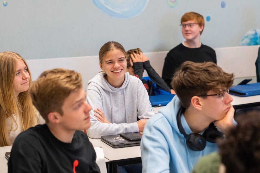 Zes jongeren zitten in een klaslokaal, twee aan twee. Op tafel liggen hun laptops.