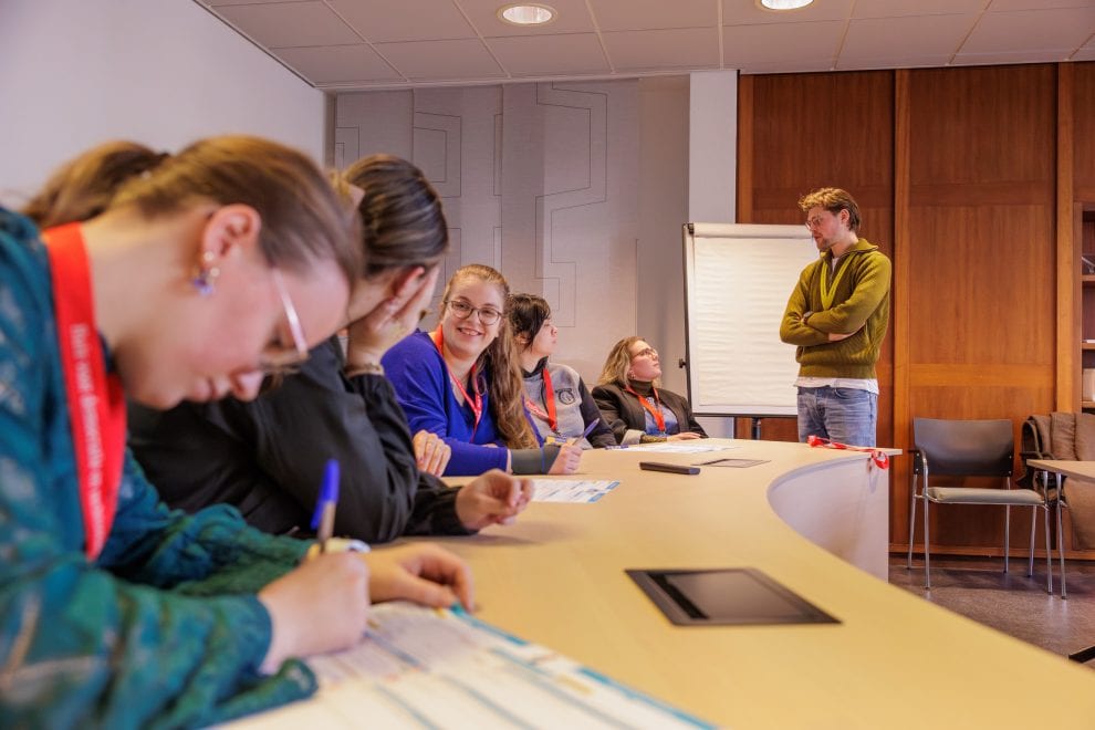 Vijf jonge vrouwen met rode keycords zitten in een leslokaal op werkbladen te schrijven. Eén vrouw praat met een jongeman die naast de tafel staat. Hij draagt een geel keycord.
