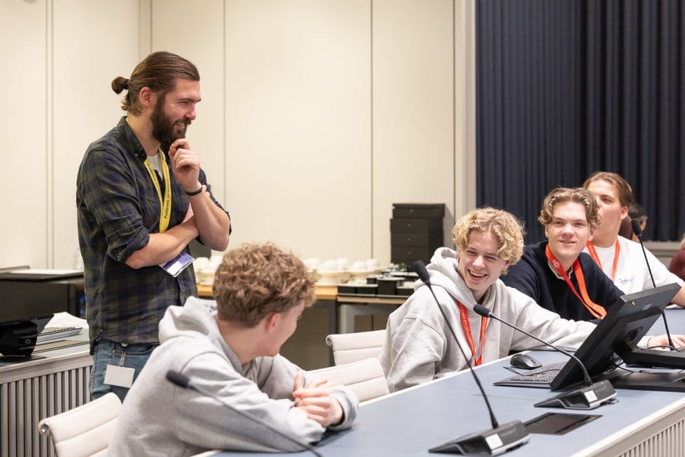 Vier jongens zitten lachend achter een tafel met microfoons. Een man staat lachend bij hen.