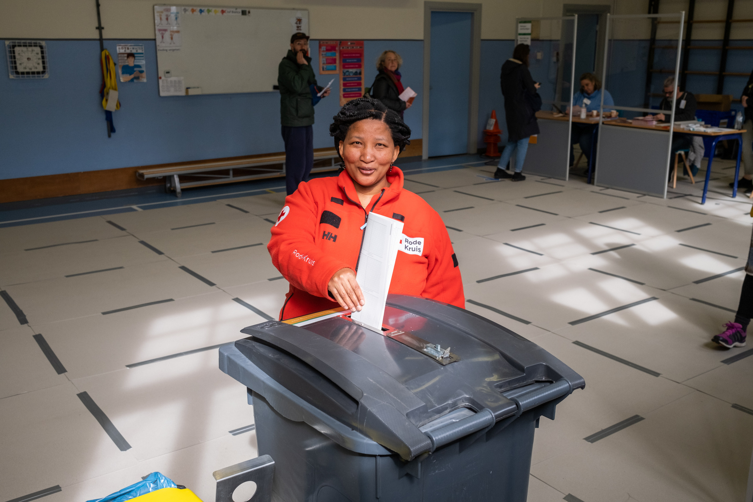 Op de foto doet een mevrouw in een rode jas een stembiljet in de stembus. Op de achtergrond zien we mensen in de rij staan voor een tafel met stembureauleden. De stembureauleden controleren de stempas van mensen die willen stemmen.