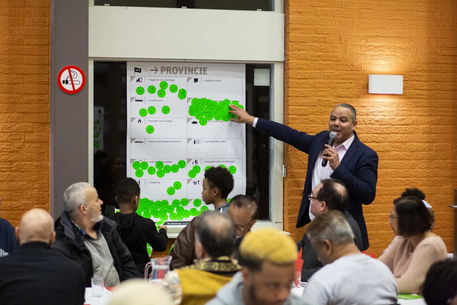 Op de foto zien we een man voor een bord met stickers staan. Boven de stickers staat het woord 'Provincie'. Hij geeft uitleg en wijst naar het bord. In de zaal zitten mensen te luisteren.